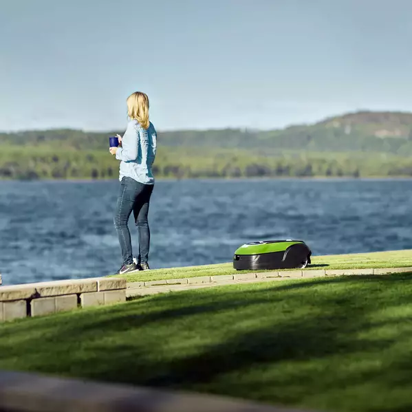 Kvinna står vid vattnet bredvid en Greenworks Optimow Robotgräsklippare